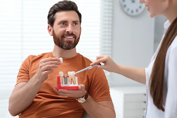 A patient holds a model of teeth with a single dental implant, while discussing recovery time of implants with his dentist.